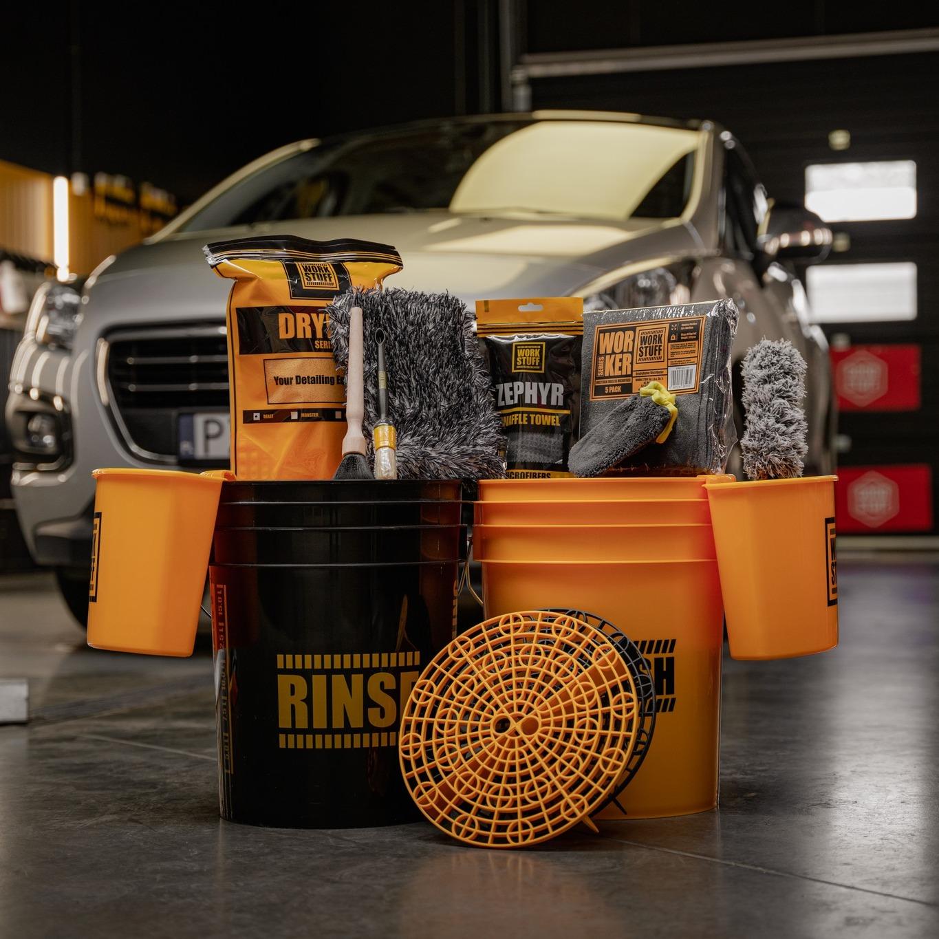 Microfiber towel, orange buckets, brushes, and cleaning products arranged before a silver SUV in a garage.