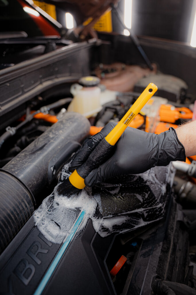 Una mano enguantada sujeta un cepillo amarillo que frota espuma limpiadora en las zonas detalladas del compartimento del motor de un coche. Una mano enguantada sujeta un cepillo amarillo que frota espuma limpiadora en las zonas detalladas del compartimento del motor de un coche.