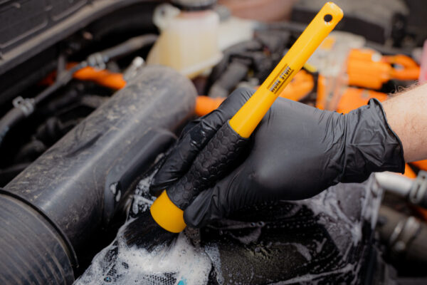 Une personne portant des gants noirs utilise une brosse jaune et du savon pour nettoyer le moteur d'une voiture, créant ainsi de la mousse sur le capot du moteur. Une personne portant des gants noirs utilise une brosse jaune et du savon pour nettoyer le moteur d'une voiture, créant ainsi de la mousse sur le capot du moteur.
