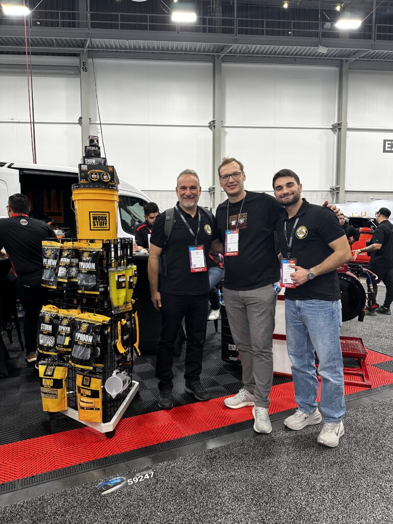 Three men in black shirts with name badges smile at a SEMA Show 2025 booth displaying car care products and bottles. Three men in black shirts with name badges smile at a SEMA Show 2025 booth displaying car care products and bottles.