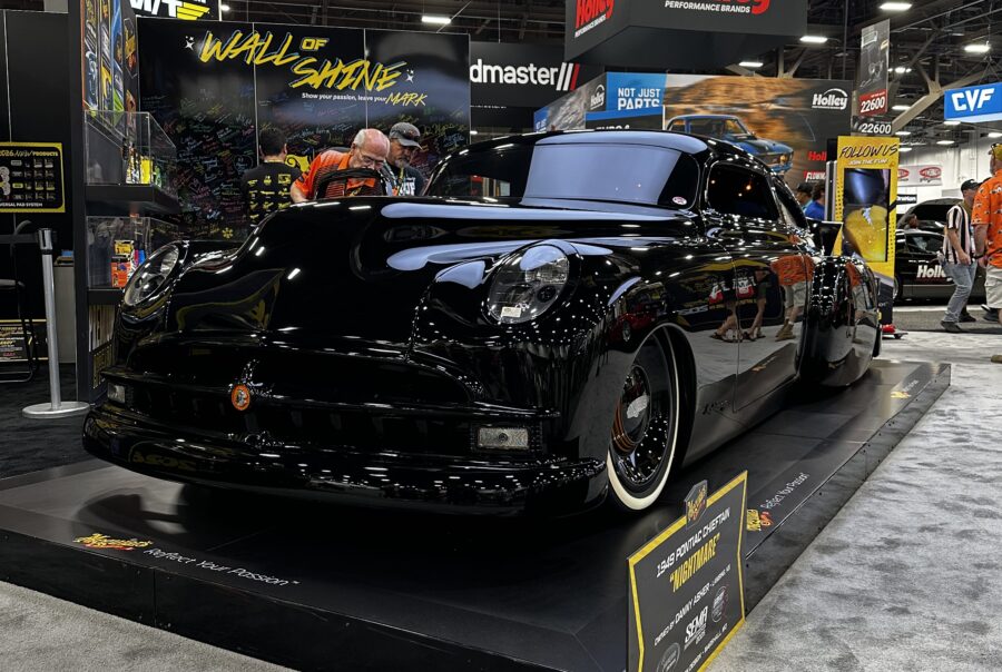 A shiny black classic car on a carpeted platform at SEMA Show 2025, with Holley above and Wall of Shine signage behind.