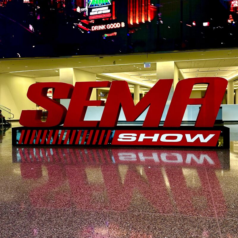 Duży czerwony napis "SEMA SHOW" znajduje się na błyszczącej, nakrapianej podłodze z kolorowymi cyfrowymi ekranami i odbiciami za nim.