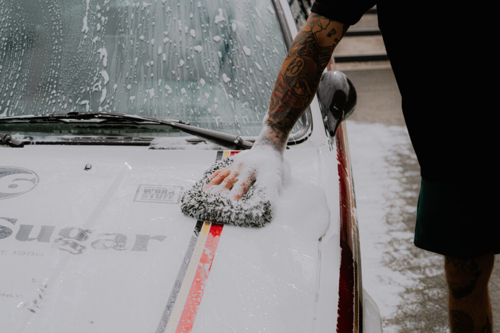 Una persona tatuada utiliza jabón y una esponja para lavar un coche blanco con rayas rojas y negras. Una persona tatuada utiliza jabón y una esponja para lavar un coche blanco con rayas rojas y negras.
