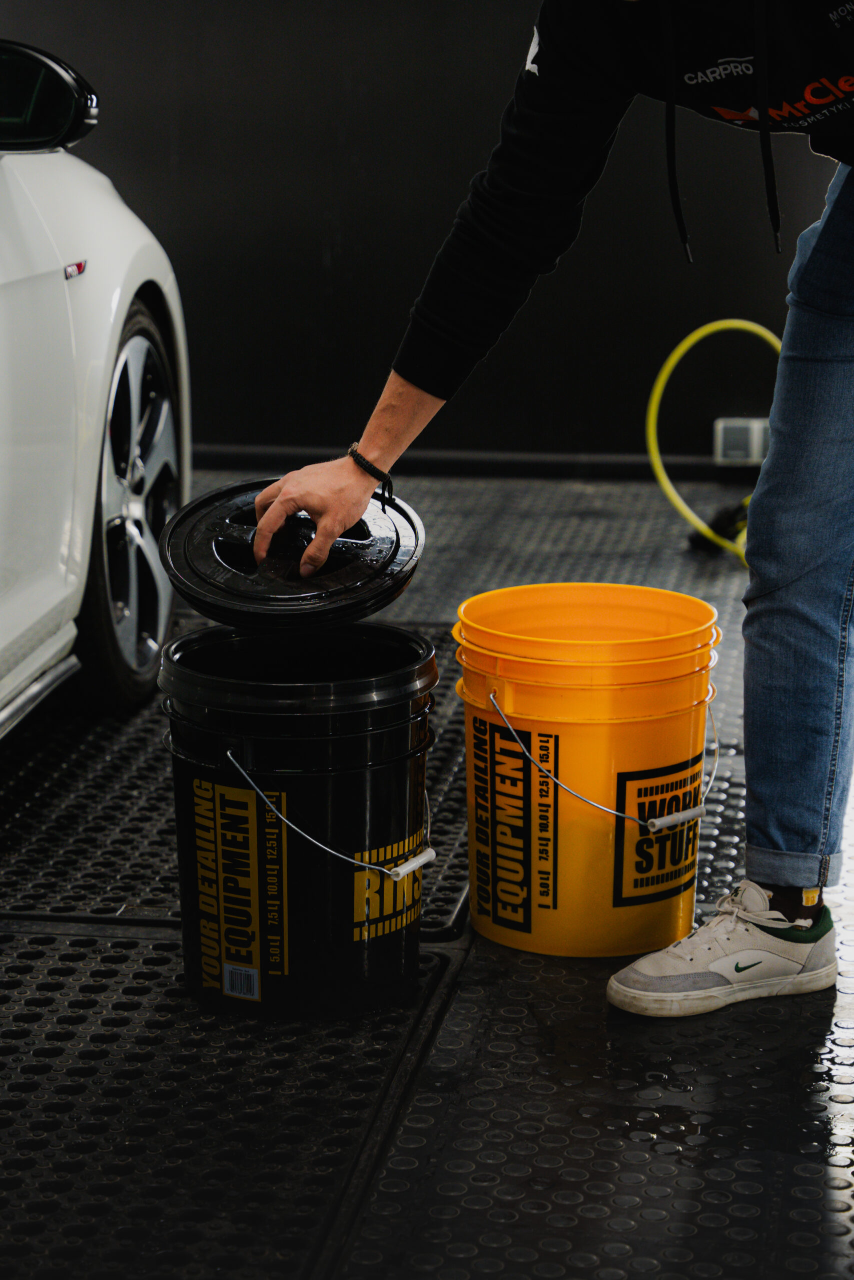 A person in jeans and sneakers lifts the HDPE lid off a black detailing bucket beside a yellow one in a car wash area.