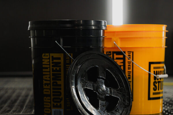 Un seau de lavage Work Stuff noir avec couvercle en forme de seau est penché vers l'avant sur un sol sombre ; un seau jaune et une lumière verticale se trouvent derrière.