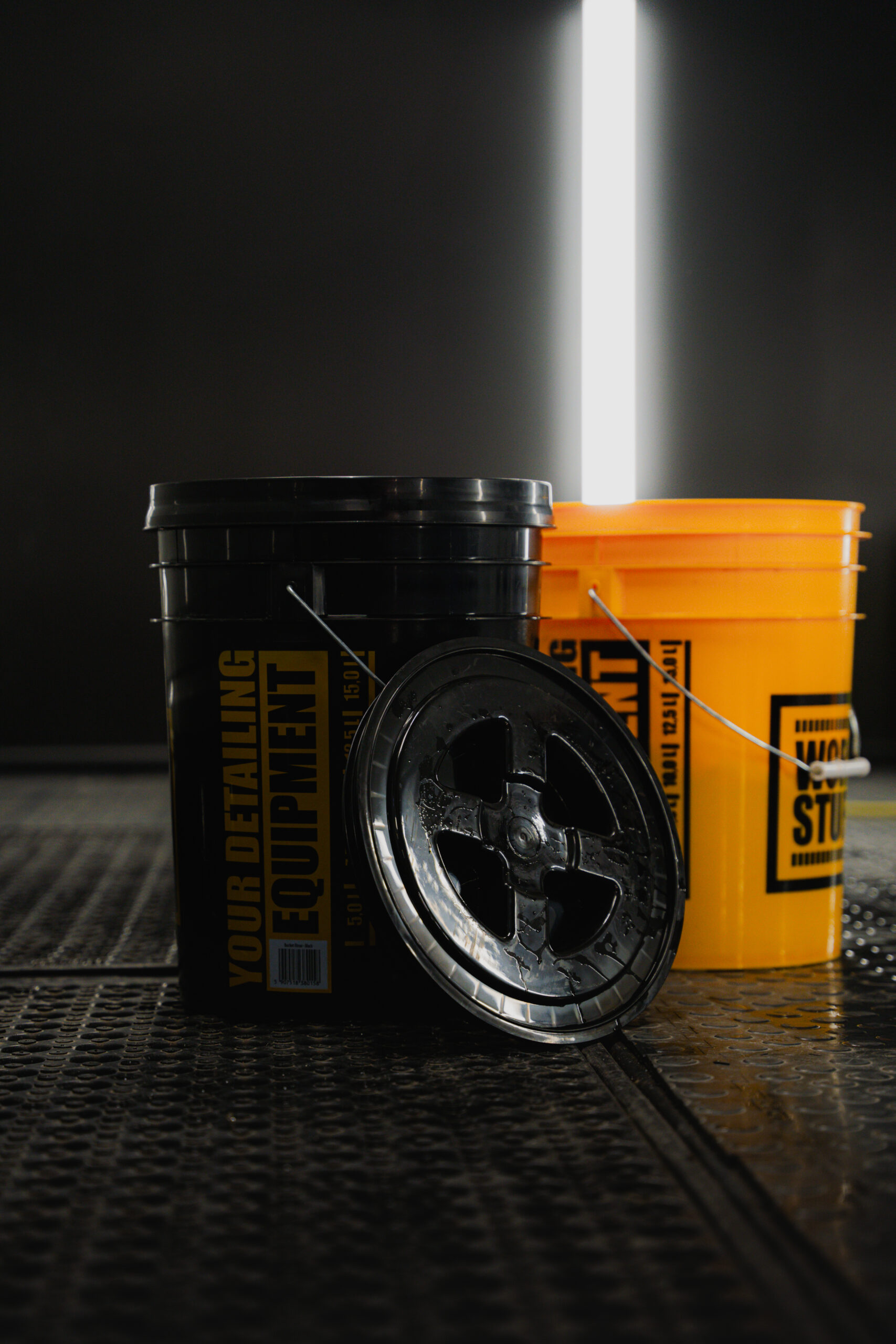 Two large buckets, one black and one yellow, labeled Work Stuff, on a dark textured floor with bright vertical backlight.