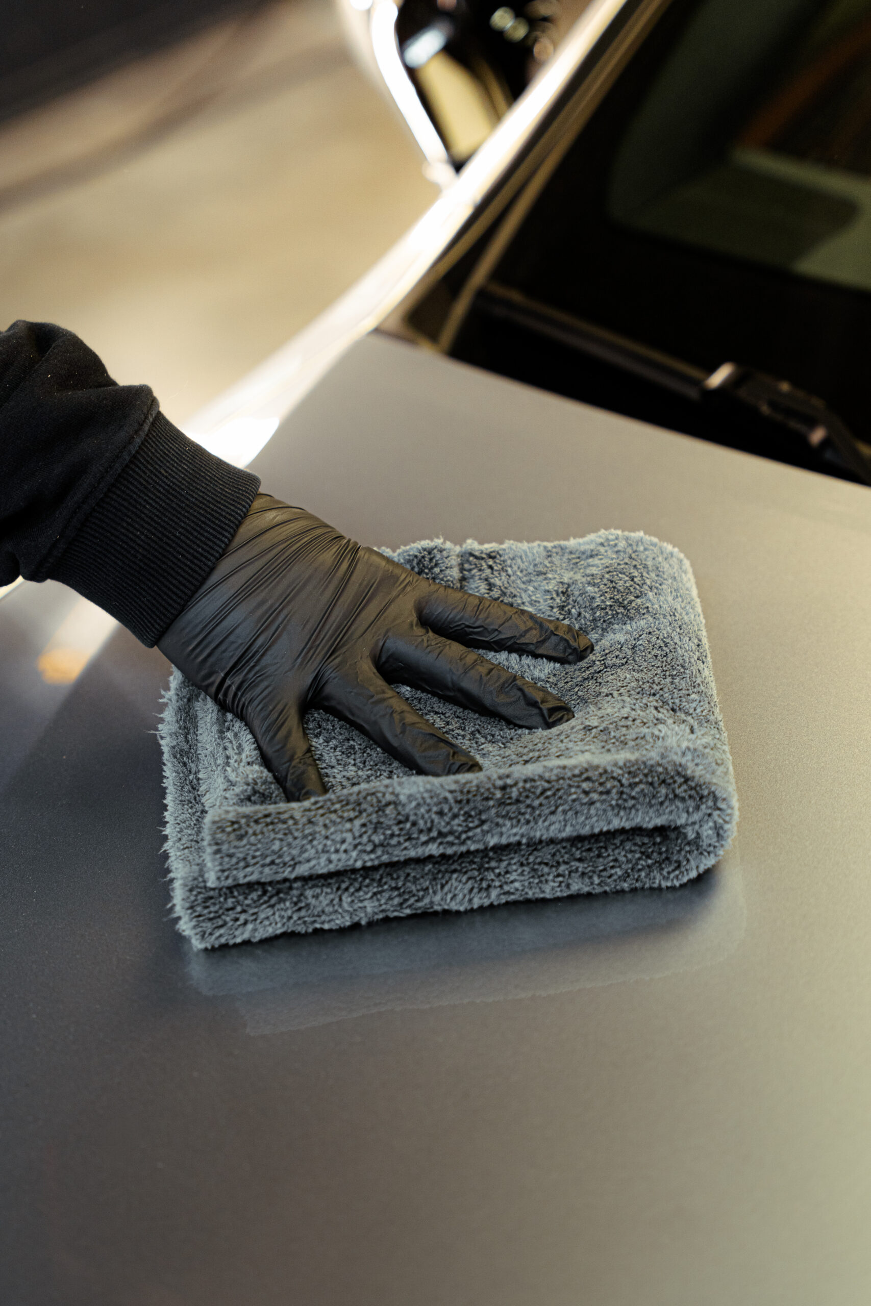A GENTLEMAN+ in a black glove carefully wipes a car’s surface with a folded gray microfiber cloth.