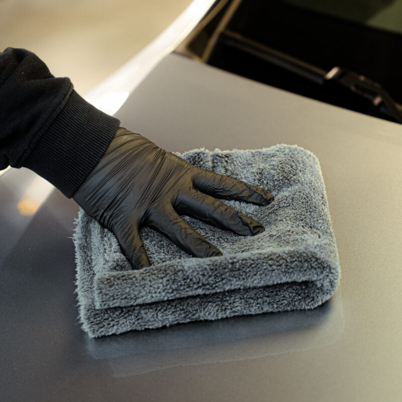 Une personne portant un gant noir essuie la surface d'une voiture avec un chiffon d'entretien en microfibres gris Work Stuff GENTLEMAN plié.