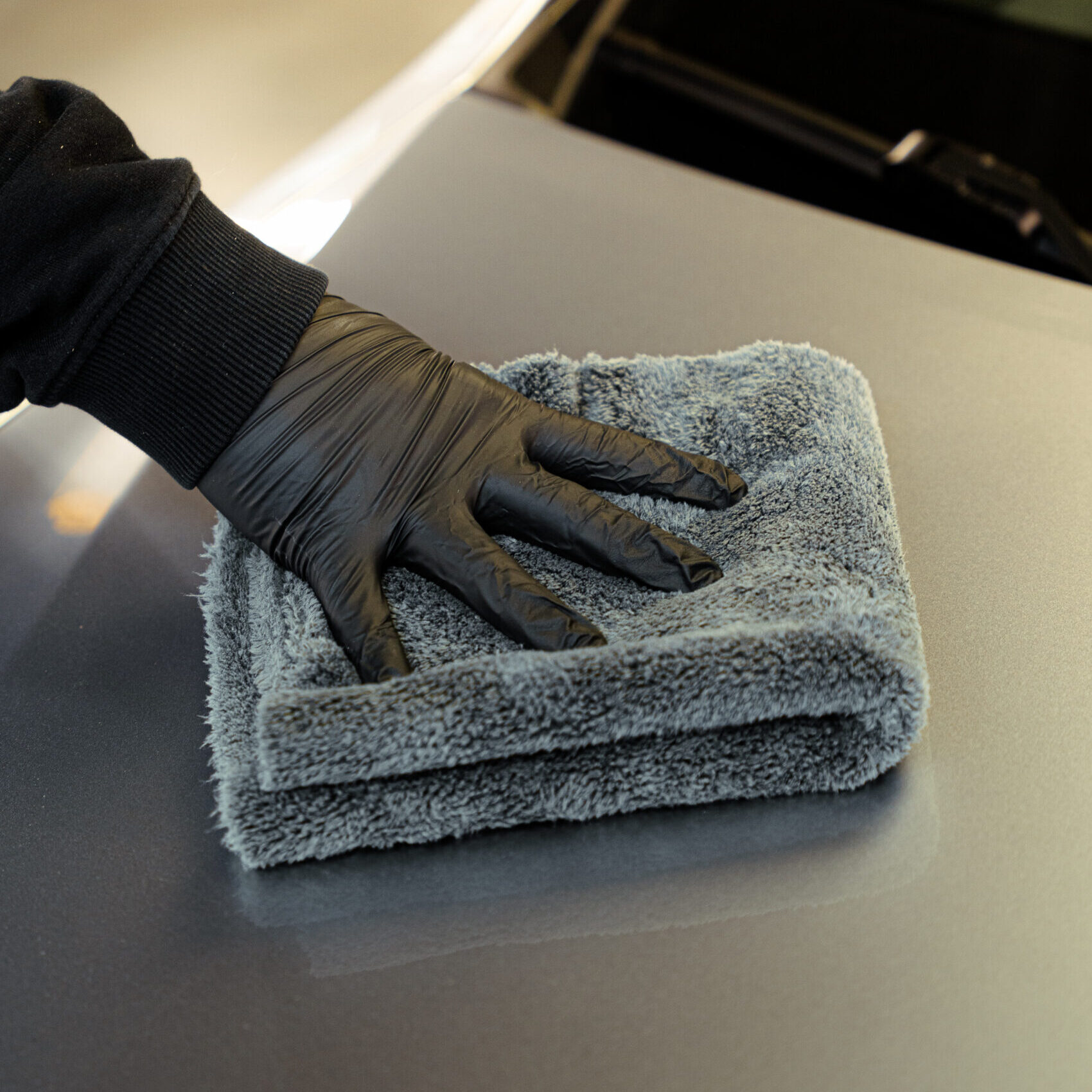 Persona con guante negro limpia la superficie de un coche con un paño gris de microfibra doblado, dejando brillo.