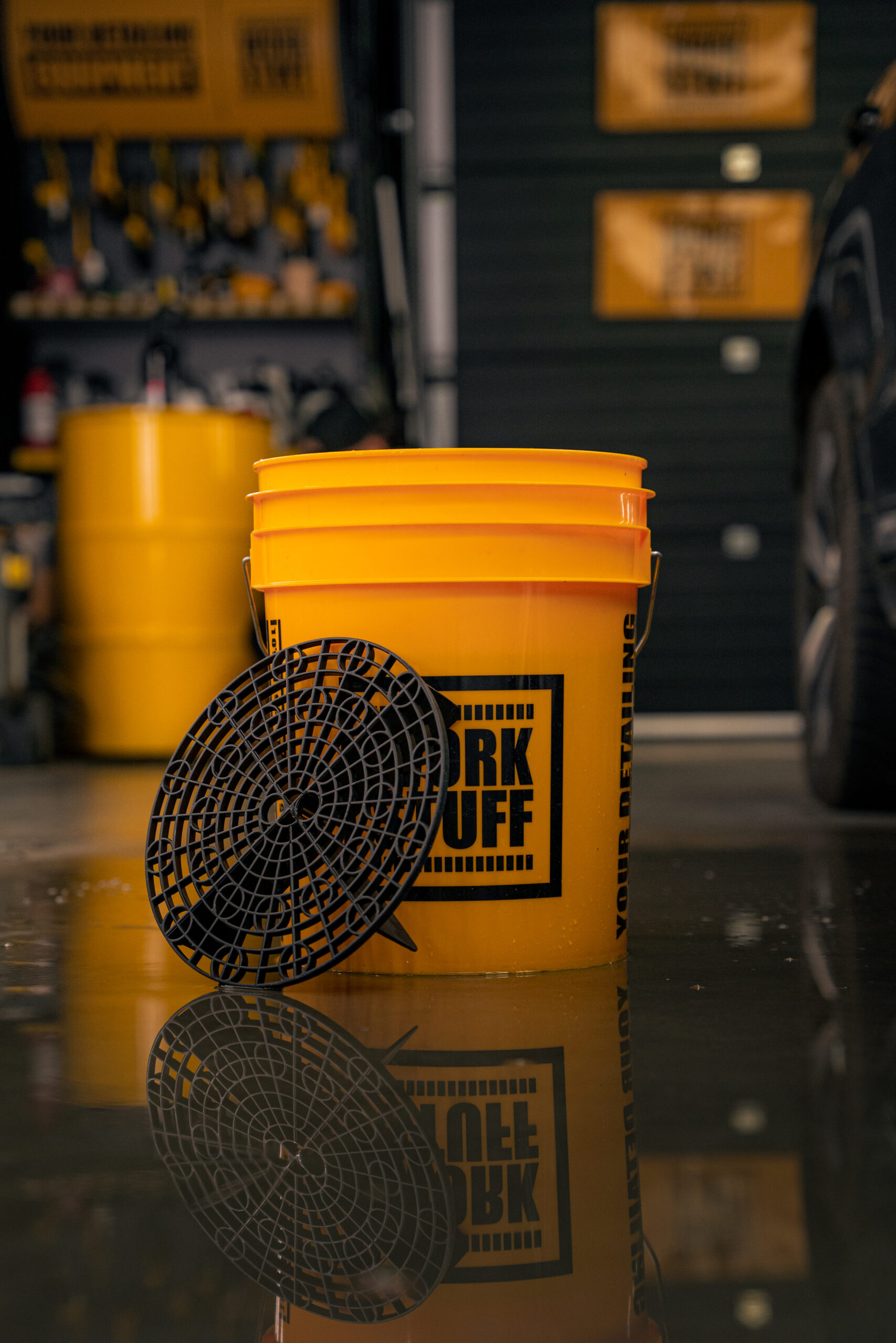 A bright yellow detailing bucket with black dirt separator sits on shiny garage floor, with yellow barrels and tools behind.