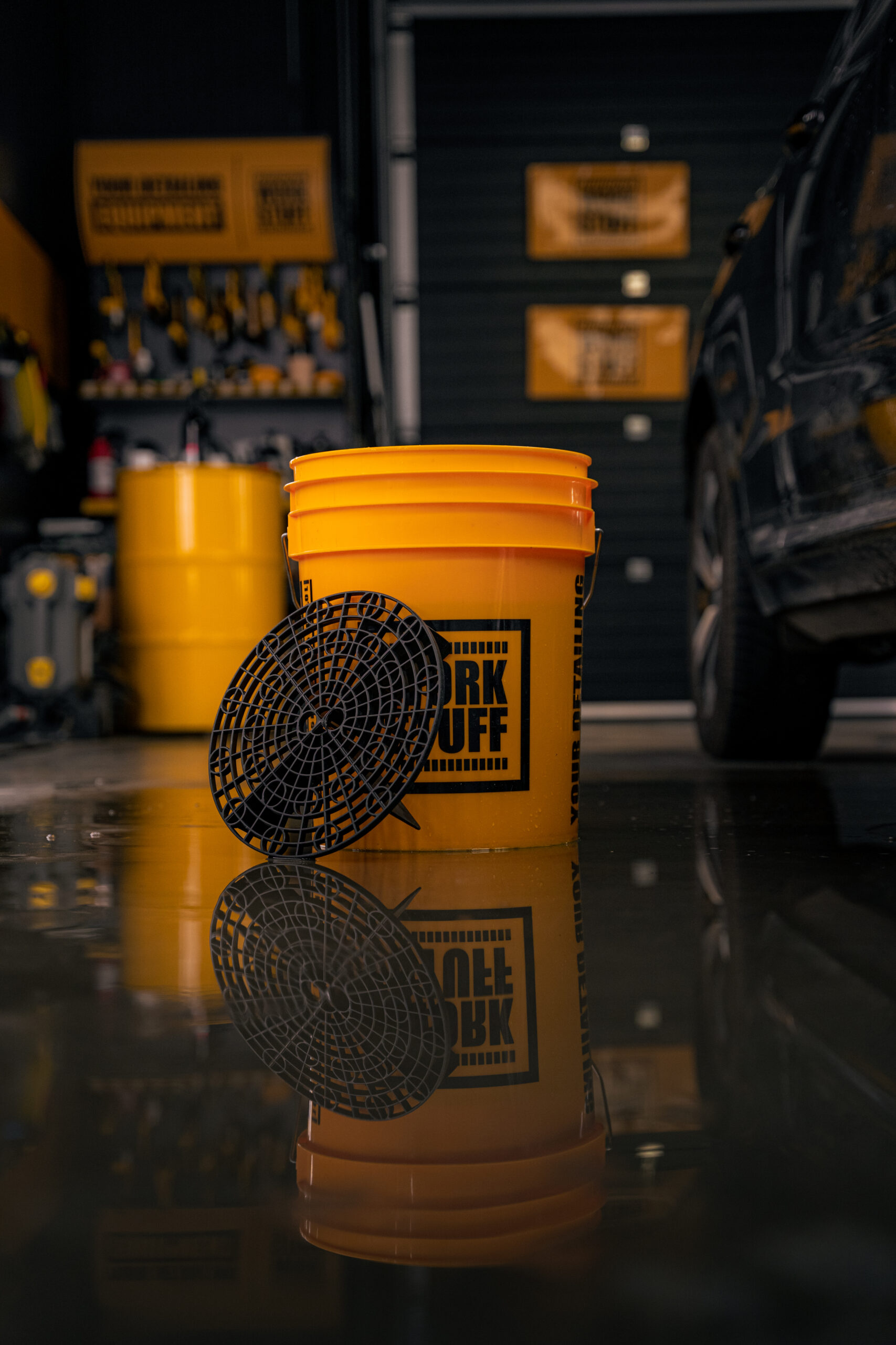 A bright yellow Work Stuff bucket with a black dirt separator on a wet garage floor, car tools and black vehicle behind.