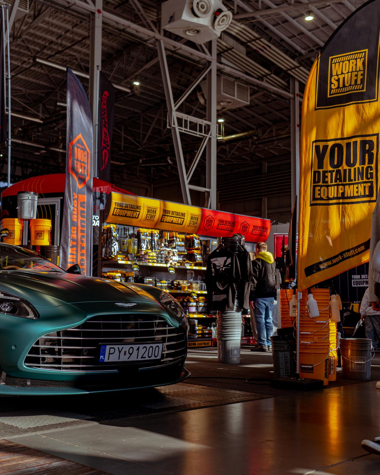 Une voiture vert mat lors d'un salon automobile intérieur à côté de bannières jaunes de Work Stuff ; des personnes recherchent des produits d'esthétique à proximité.
