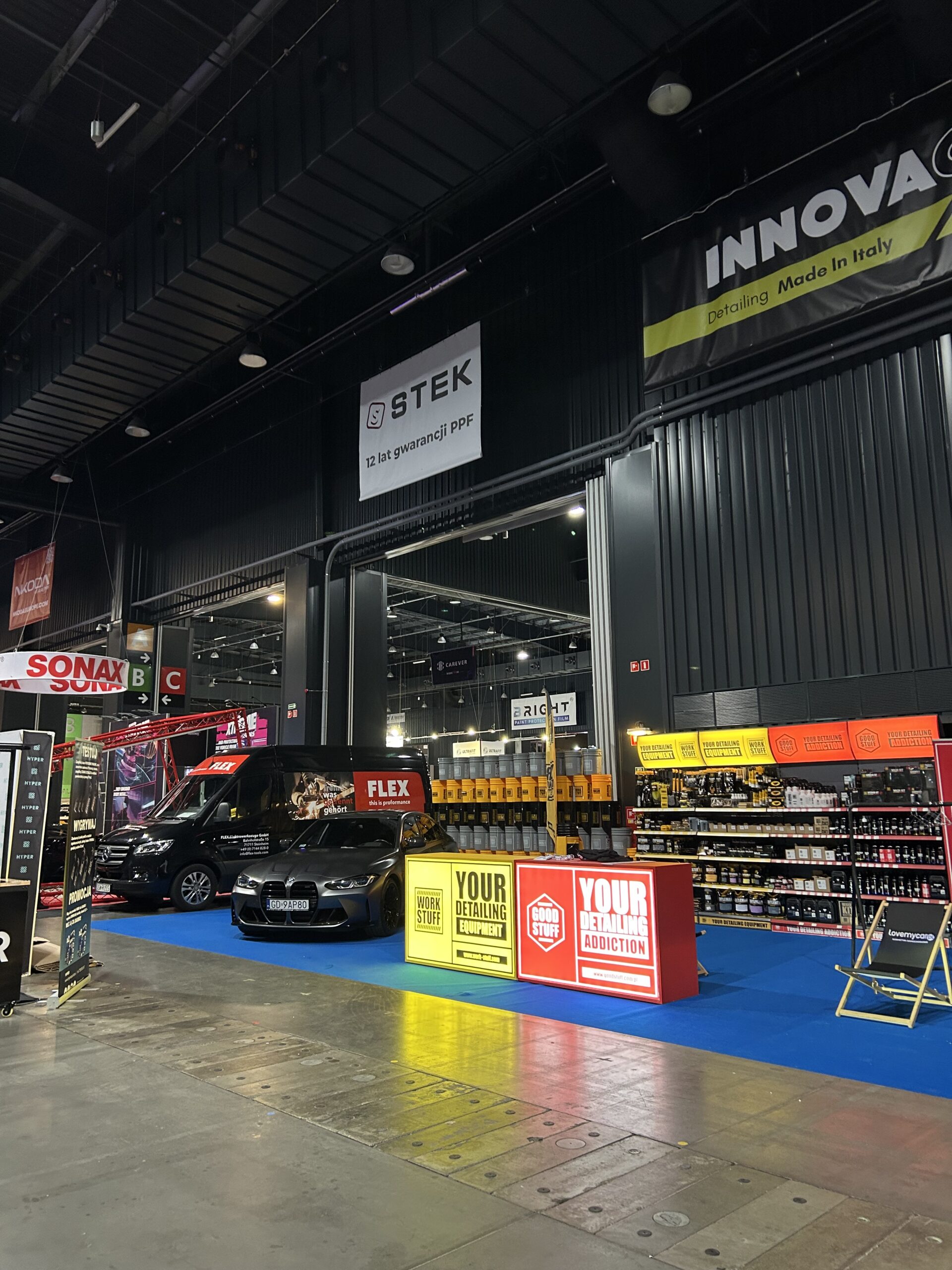 Stand d'esthétique automobile avec des bannières jaunes et rouges, des produits d'entretien automobile, une voiture noire et des panneaux de Work Stuff dans le hall.
