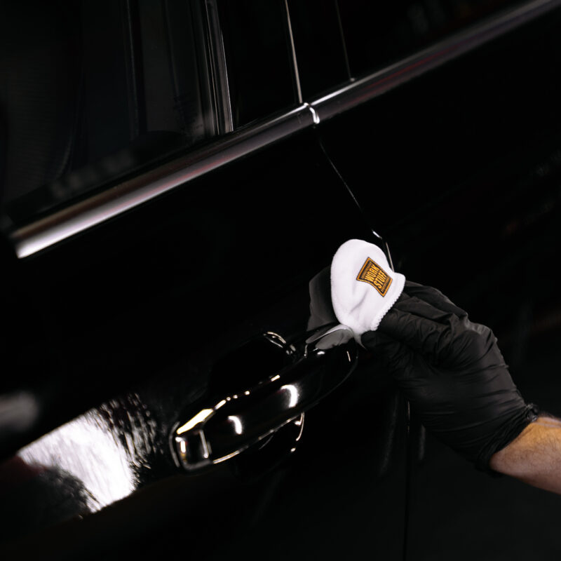 A person wearing a black glove uses a white cloth to clean or polish the handle of a black car.