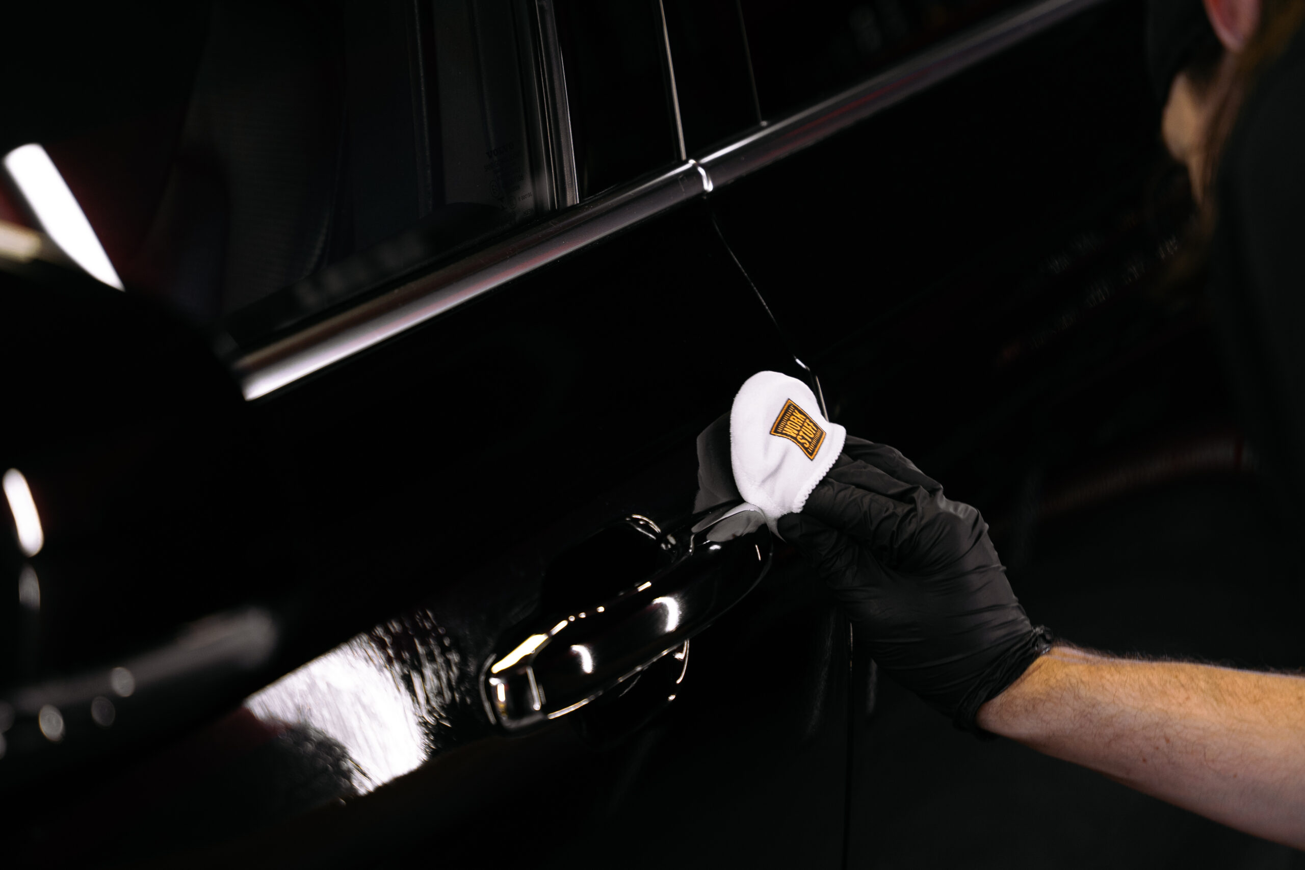 A person wearing a black glove uses a white cloth to clean or polish the handle of a black car.